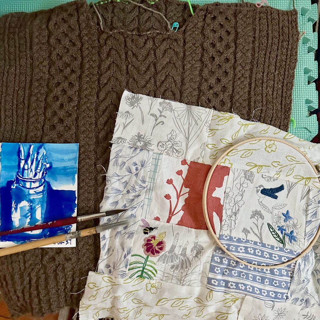creative projects flat lay: cable knit sweater pieces with a bright blue ink painting, paintbrushes, and patchwork sewing and embroidery laid out over it