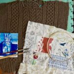creative projects flat lay: cable knit sweater pieces with a bright blue ink painting, paintbrushes, and patchwork sewing and embroidery laid out over it